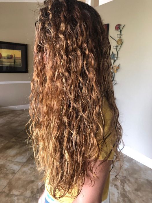 A person with long, curly hair stands indoors, facing sideways. The background features a framed picture and wall decor