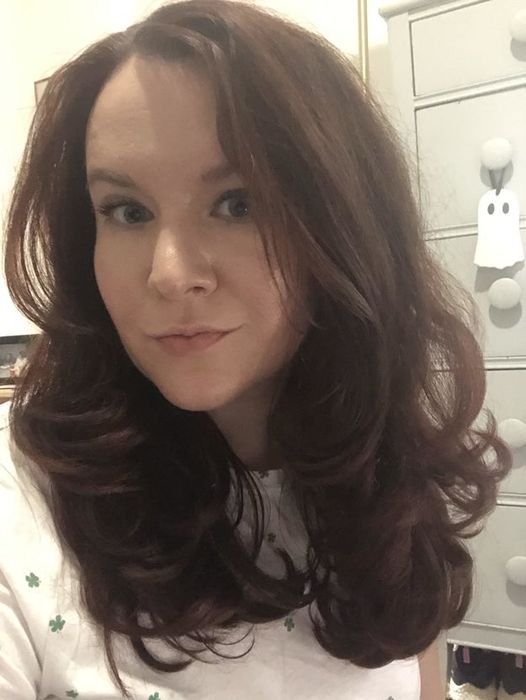 Elizabeth Lilly, with shoulder-length wavy hair, wears a light-patterned shirt and stands in front of a dresser adorned with a ghost decoration. Names of others are unknown