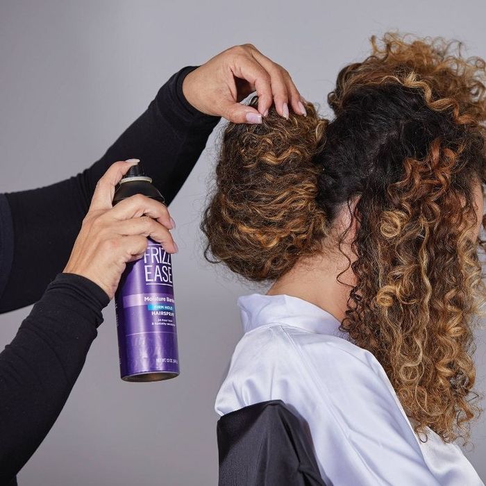 A person spraying hairspray onto a person with blonde curly hair.