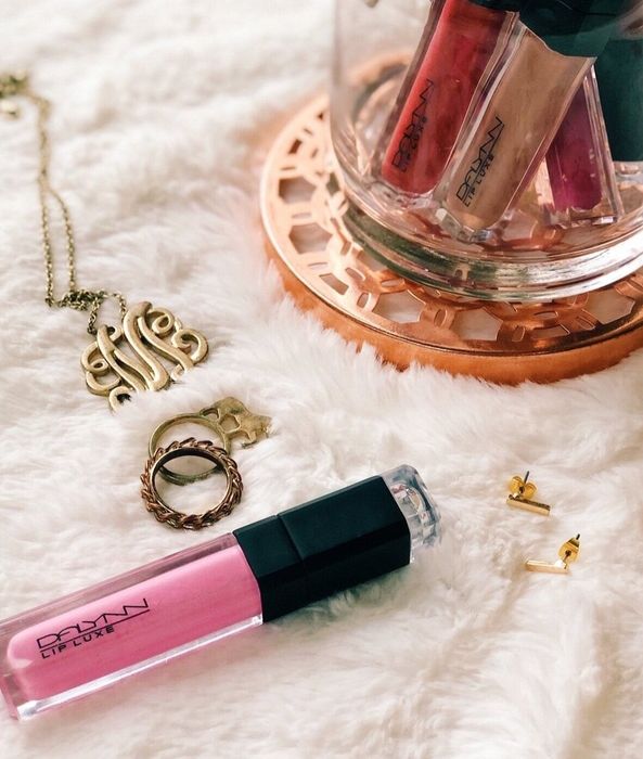 A pink and black tube of lip gloss placed next to a jar of products in various shades and some gold jewelry.