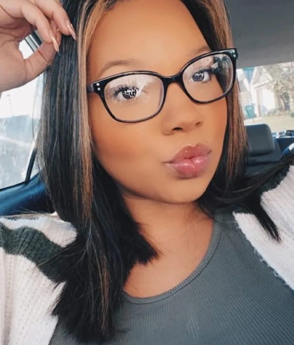 A person in a car, wearing glasses and a striped sweater, puckering their lips for a selfie.