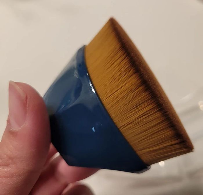 A hand holding a flat-tipped makeup brush with a blue handle.