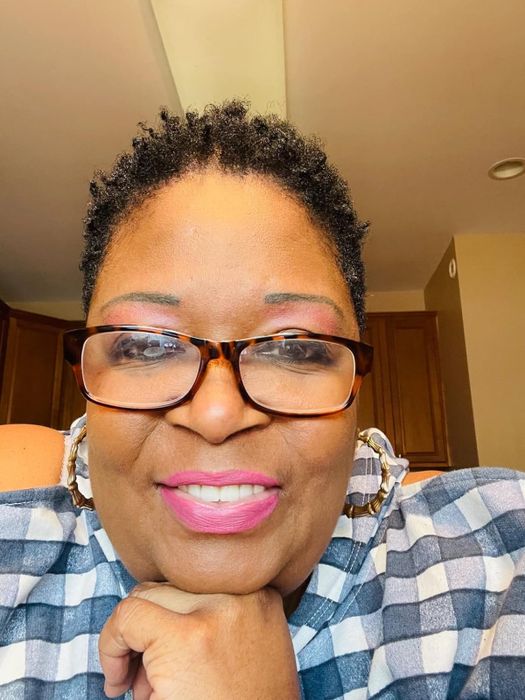 A close-up of a smiling reviewer wearing bold pink lipstick, glasses, hoop earrings, and a checkered outfit, resting her chin on her hand.