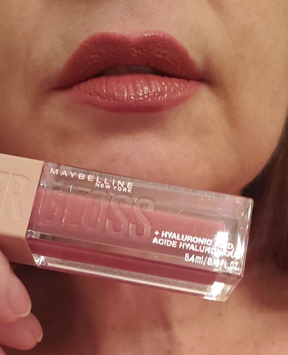 A reviewer holding a dark pink tube of lip gloss just below their lips.