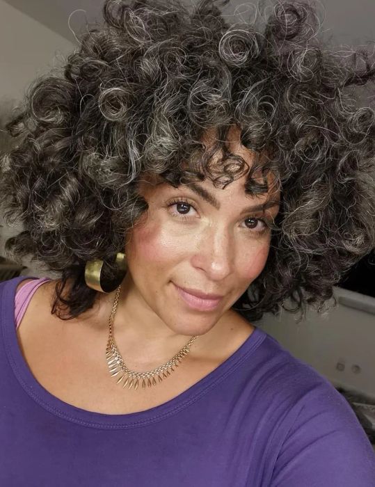 A reviewer with full, curly hair and hoop earrings, accessorized with a necklace and wearing a purple top