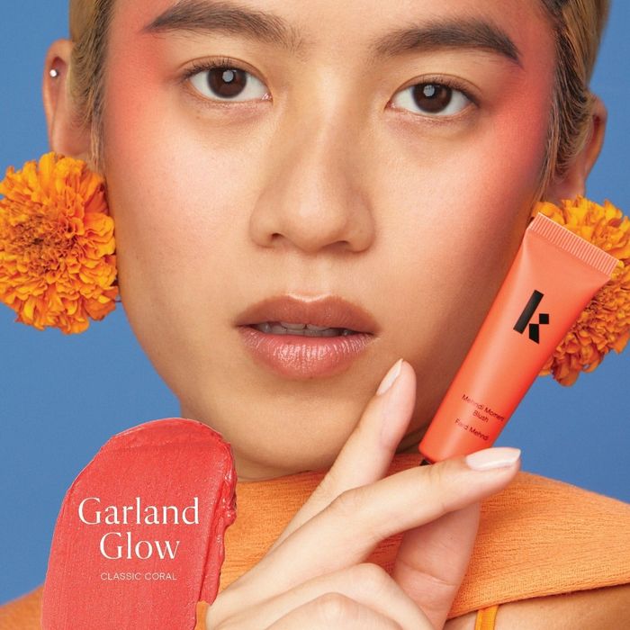 A close-up shot of a model showcasing Kaja's Garland Glow Classic Coral makeup, adorned with floral accents behind her ears and a vibrant coral blush on her cheeks.
