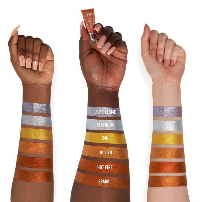 Three arms with varying skin tones displaying six swatches of foiled liquid eyeshadow in shades labeled: Lilac Flame, Platinum, 24K, Gilded, Hot Fire, and Spark.