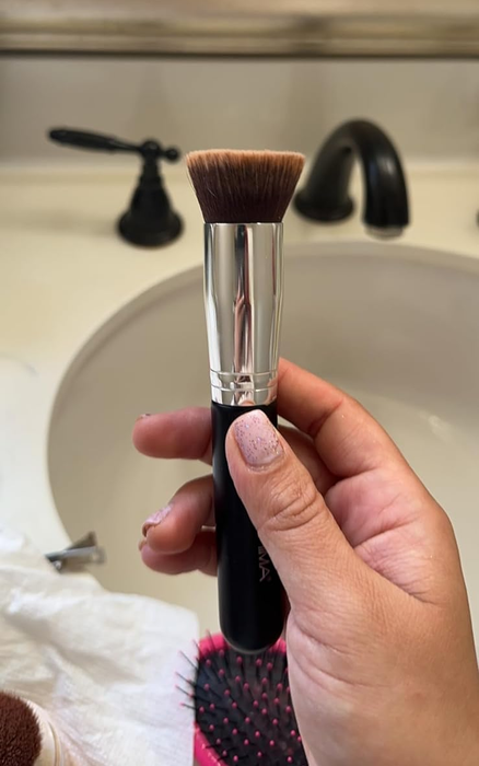 A reviewer holding a makeup brush in front of a bathroom sink and mirror, with another person applying makeup visible in the reflection.