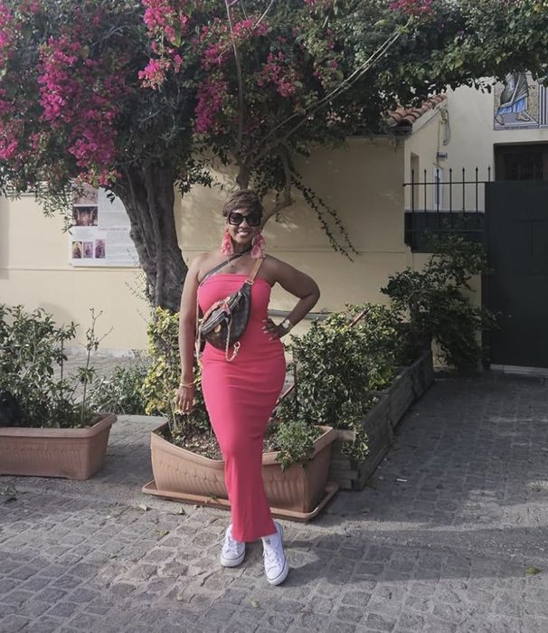 reviewer in a pink sleeveless dress striking a pose with one hand on her hip, accessorized with a belt and sunglasses, standing near blooming plants