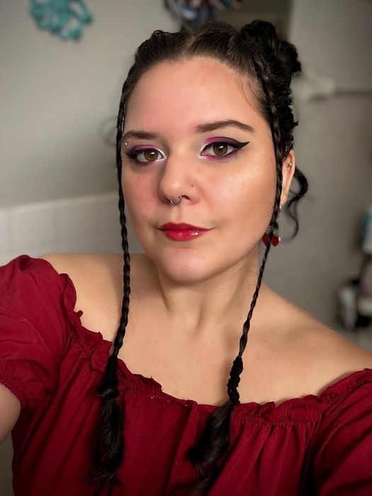 A reviewer with braided hair, wearing a red off-the-shoulder top, poses for a selfie. Her look includes winged eyeliner, red lip gloss, and a nose piercing.