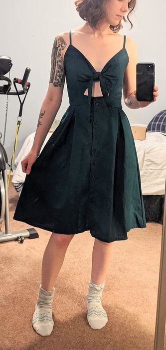Person in a sleeveless dress featuring a front tie, capturing a mirror selfie. Tattoos visible on the left arm