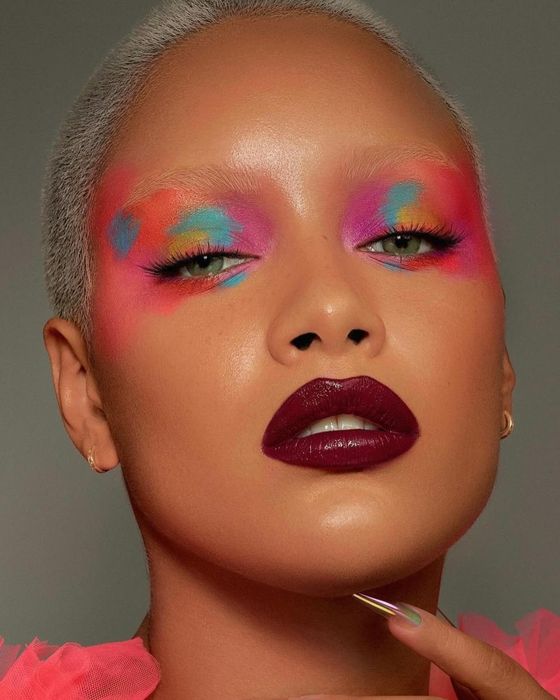 A close-up of a woman showcasing bold, artistic eye makeup, dark lipstick, and short platinum blonde hair.