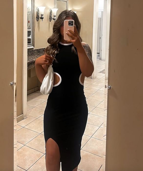 Person in a black dress featuring cutouts, holding a white bag, taking a mirror selfie