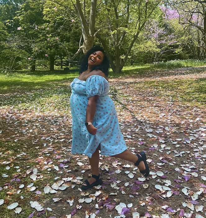 reviewer in a floral dress standing in a park surrounded by fallen petals, smiling with one hand resting on her hip