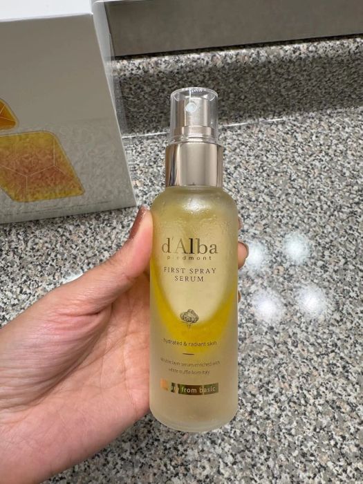 A hand holds a bottle of d'Alba Piedmont First Spray Serum on a speckled countertop.