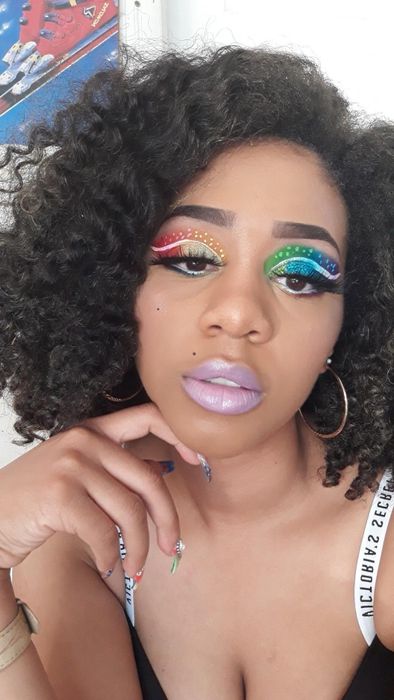 A curly-haired woman showcasing vibrant rainbow eye makeup adorned with rhinestones, paired with light lavender lipstick, hoop earrings, and a chic black-and-white top.