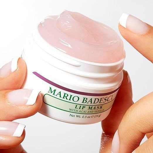 A person holding a Mario Badescu lip mask jar, with acai and vanilla in the background. The jar contains 0.5 oz (14 g), with the person's nails polished with white tips.