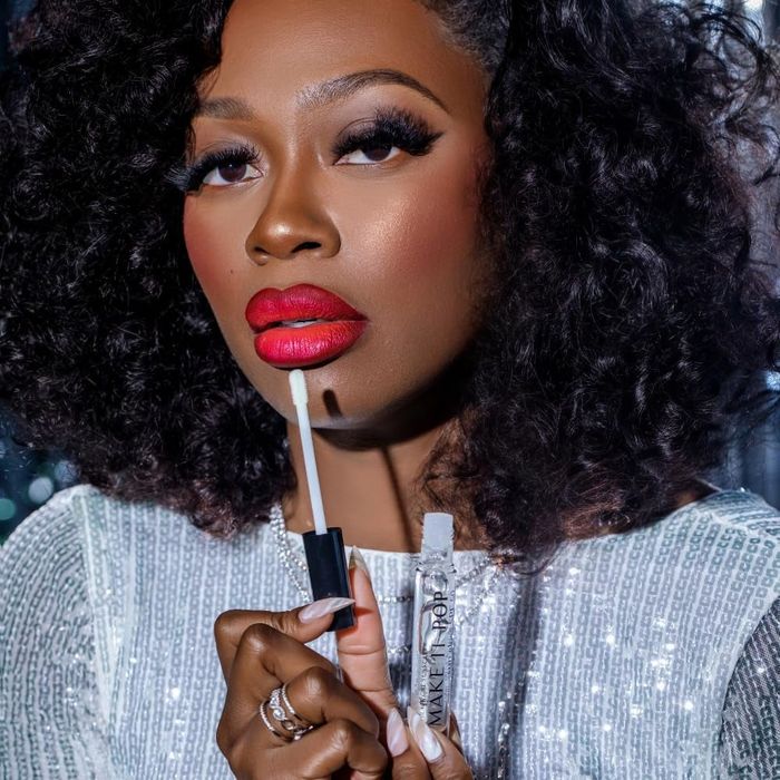A curly-haired model wearing bold red lipstick applies the Make It Pop SPF lip gloss