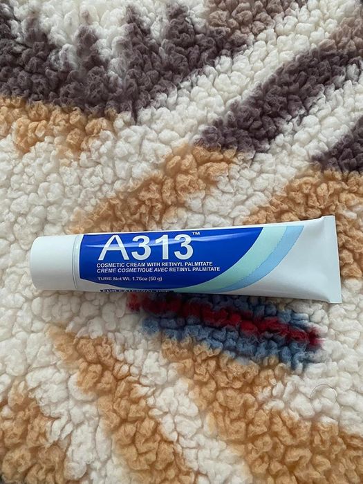 A tube of A313 cosmetic cream, featuring retinyl palmitate, placed on a textured surface. The cream comes in a white and blue container.