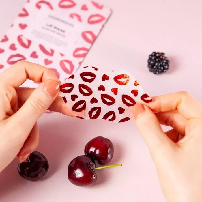 Model holding a lip and heart-printed face mask sheet over cherries and a blackberry, with product packaging visible in the background