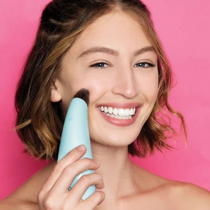 A smiling model with shoulder-length hair uses a blue device on their face against a pink background