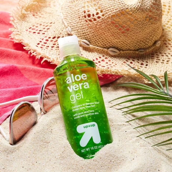 A bottle of aloe vera gel placed on a sandy beach alongside sunglasses, a straw hat, and a palm leaf
