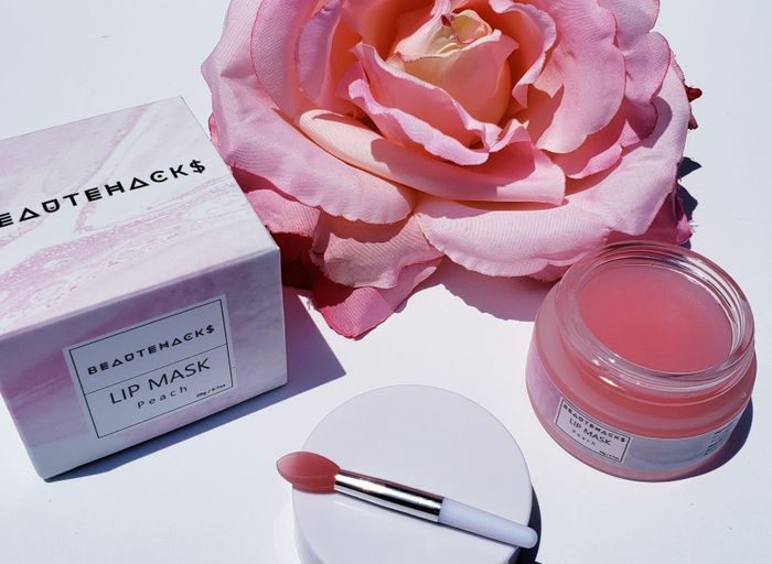 BeauteHacks lip mask set, featuring a boxed lip mask, an open jar of peach lip mask, a small applicator, and a large pink flower in the background