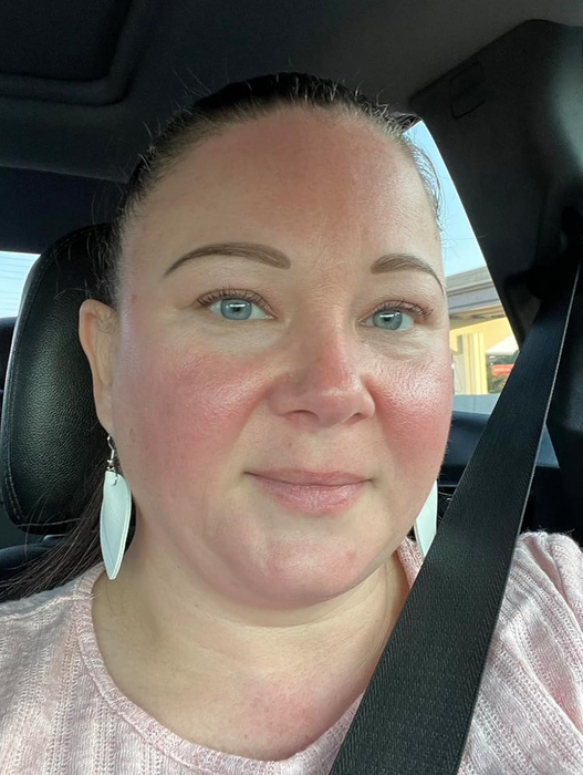 A person sitting in a car with a seatbelt on, wearing a light, textured top and teardrop-shaped white earrings.