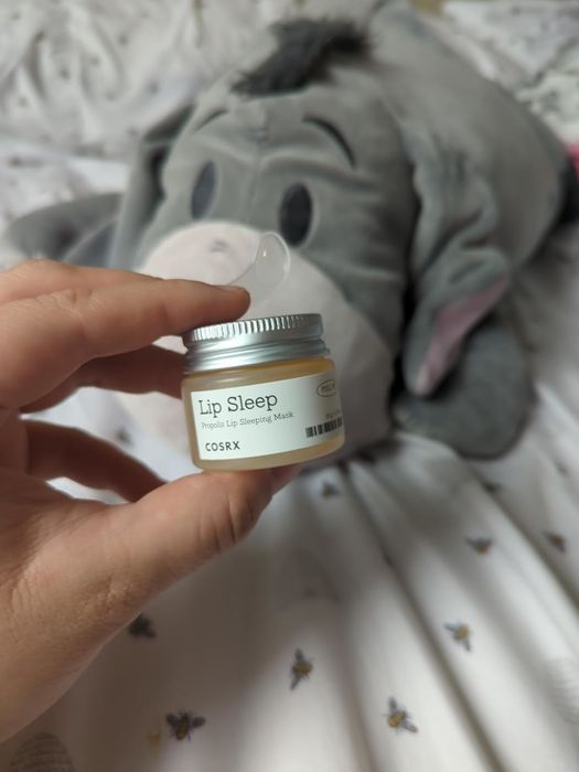 Hand holding a jar of COSRX Lip Sleep mask in front of a plush donkey toy placed on a bed.