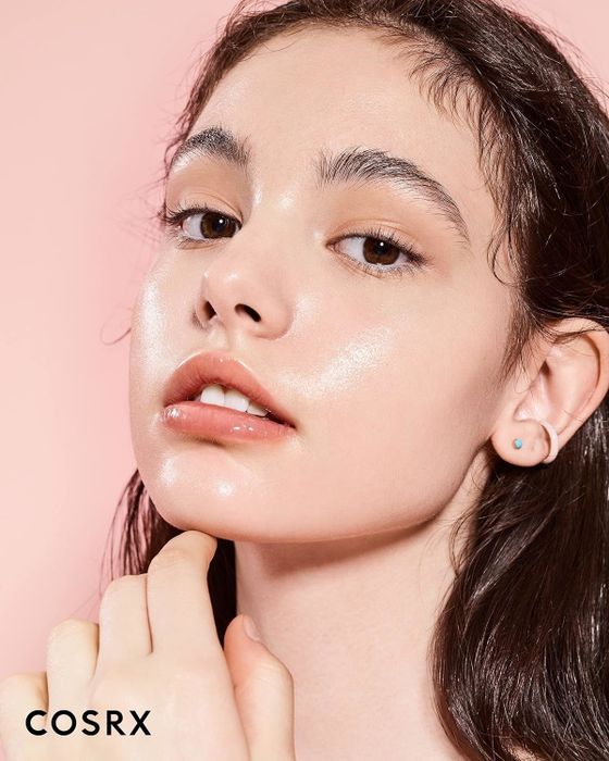 Woman with glowing, dewy skin gently touching her chin, promoting COSRX skincare product.
