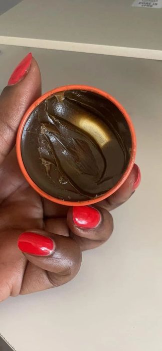 A reviewer's hand with red nails holding an open container of brown shea butter