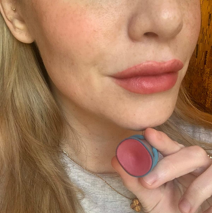A person holding a round container of pink lip balm near their lips, which are lightly tinted with the product