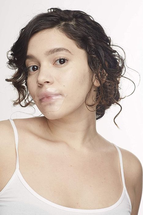 A person with short curly hair applies a transparent, adhesive face mask to their lips and chin, wearing a white tank top