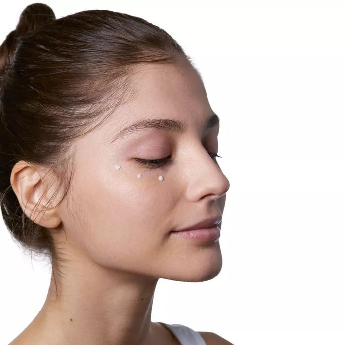 A model with a bun demonstrates applying dots of cream under the eye