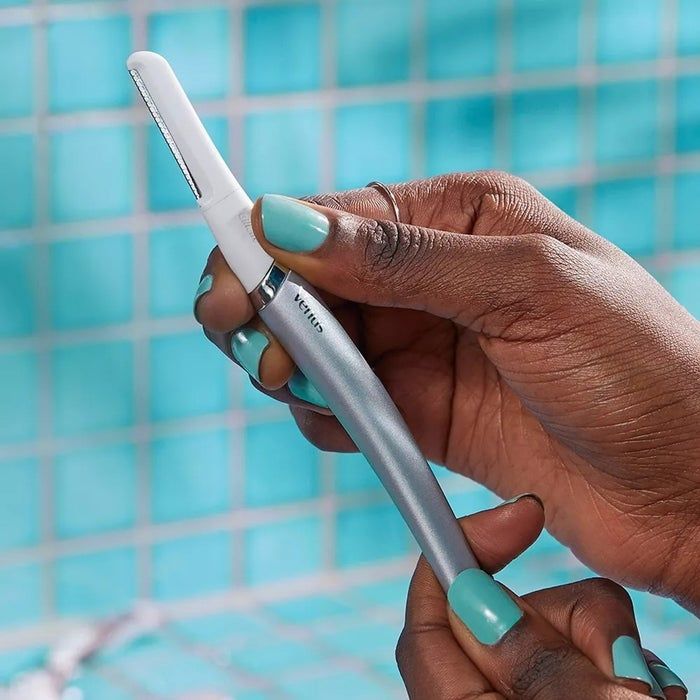 A model with perfectly manicured nails holds a Venus facial razor against a backdrop of a tiled bathroom wall