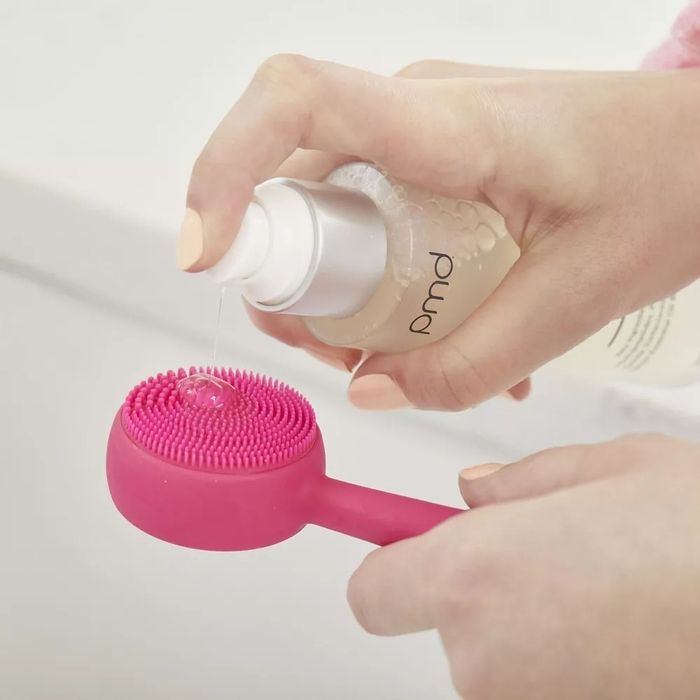 Model holding a cleanser bottle, dispensing product onto a pink facial cleansing brush