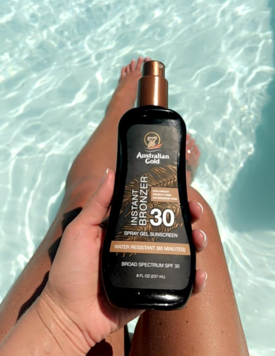 A hand holding a bottle of Australian Gold SPF 30 Instant Bronzer Spray Gel by a pool, with the label highlighting 80 minutes of water resistance