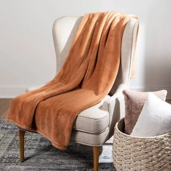 A cozy beige chair with a soft blanket draped over it, beside a basket filled with two pillows, resting on a patterned rug.