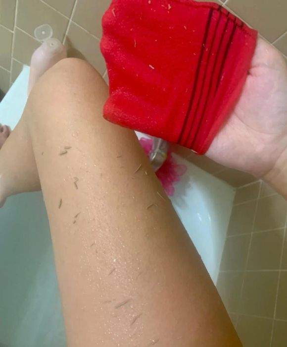 A person exfoliating their leg in a bathtub using a red exfoliating mitt.