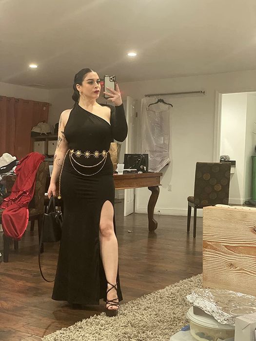 Amazon reviewer modeling a black long dress with one long sleeve and a gold belt at the waist