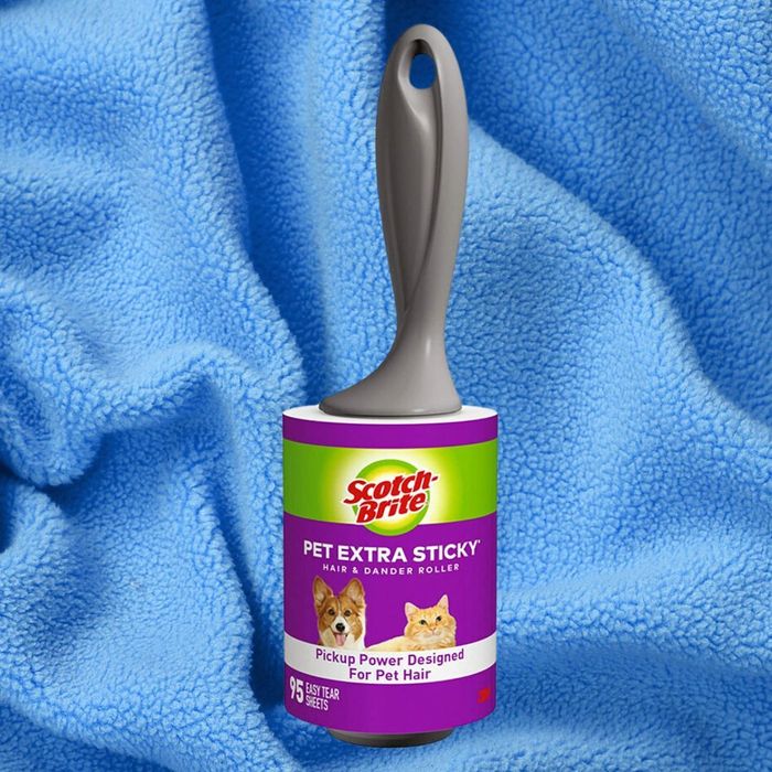 The Scotch-Brite Pet Extra Sticky lint roller is seen resting on a blue blanket, designed to effectively pick up pet hair. The label prominently features images of a dog and a cat.