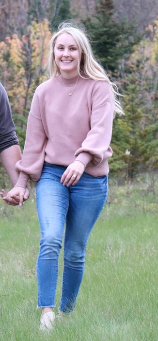Person smiling while walking outdoors, dressed in a pink sweater and blue jeans