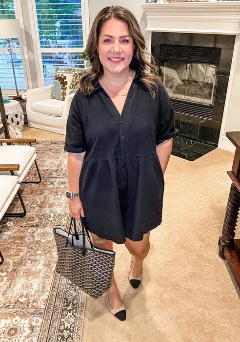 reviewer in a black dress holding a patterned tote bag, standing indoors near a fireplace