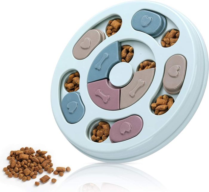A circular interactive pet puzzle toy with sliding compartments filled with kibble, accompanied by scattered kibble outside the toy for display.