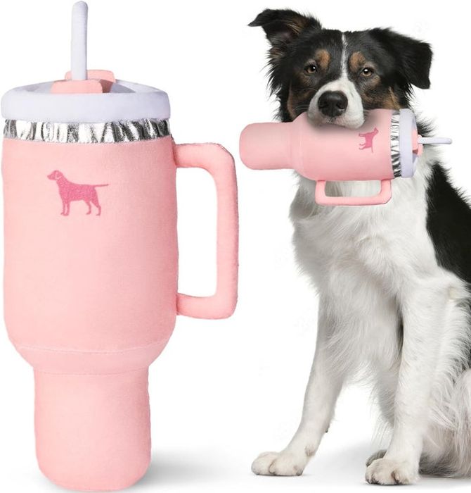 A dog holds a large, plush, pink water bottle toy in its mouth. The toy includes a handle and an embroidered dog design.