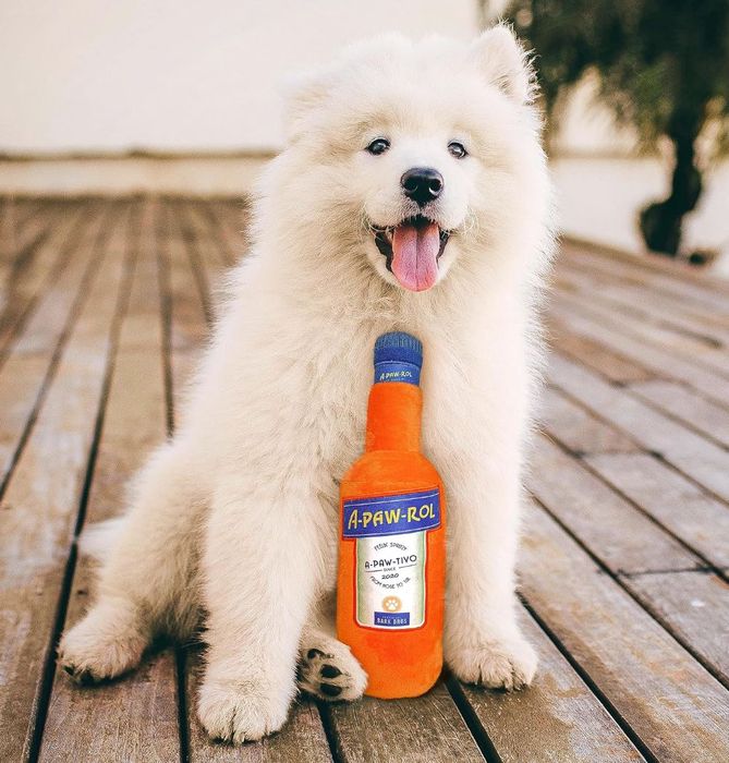 A fluffy white puppy sits on a wooden deck, joyfully holding a toy bottle labeled