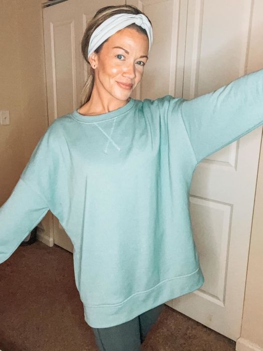 A person in an oversized sweatshirt and headband strikes a pose indoors, arms outstretched. Featured in a shopping article highlighting cozy loungewear.