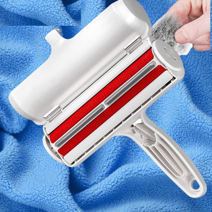 A hand grips a pet hair remover roller positioned over a blue fabric surface. The roller features a handle and a bright red cleaning area.