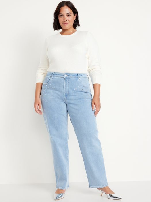 A person dressed in a white sweater and light denim jeans with floral embellishments, standing in a neutral background.