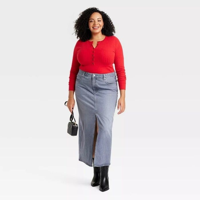 A model styled in a long gray denim skirt, paired with a red button-up top, black ankle boots, and a small handbag